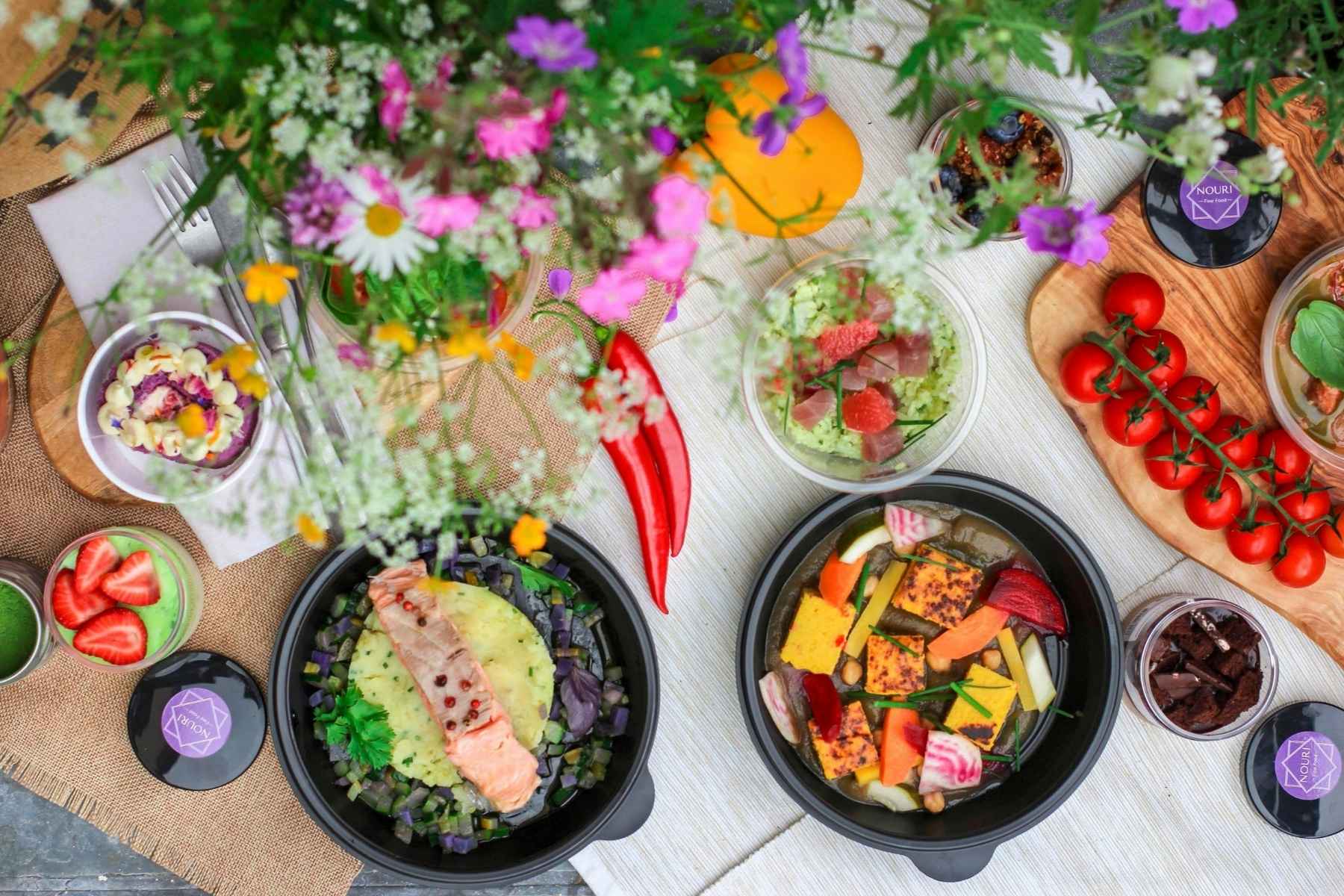A colorful picnic spread with various healthy dishes, including grilled salmon, tofu, fresh vegetables, cherry tomatoes on the vine, strawberries, and flowers arranged on a light cloth.
