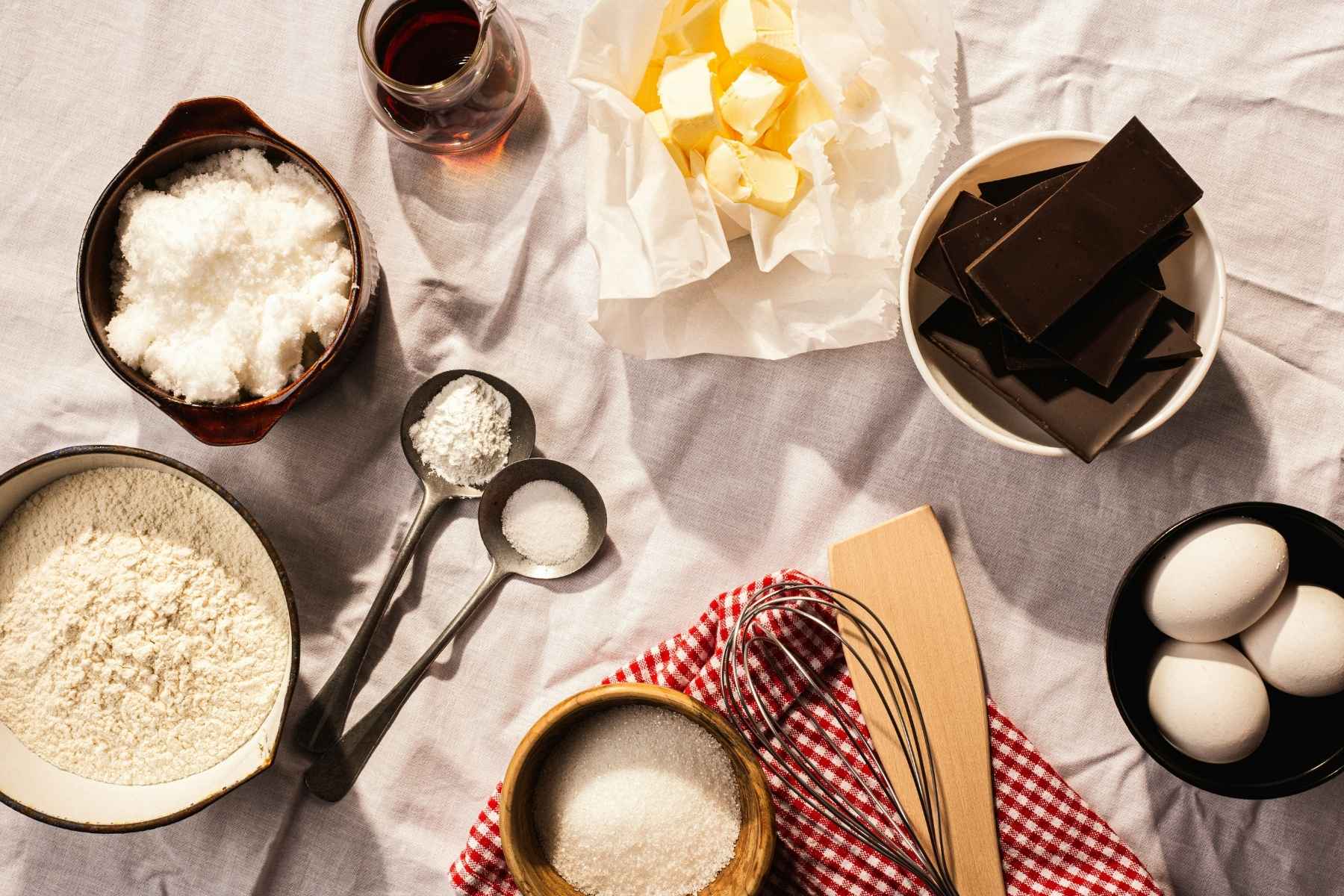 A flat lay of baking ingredients on a white cloth, including flour, sugar, shredded coconut, eggs, chocolate, butter, vanilla extract, measuring spoons, a whisk, and a wooden spatula on a red checkered napkin.