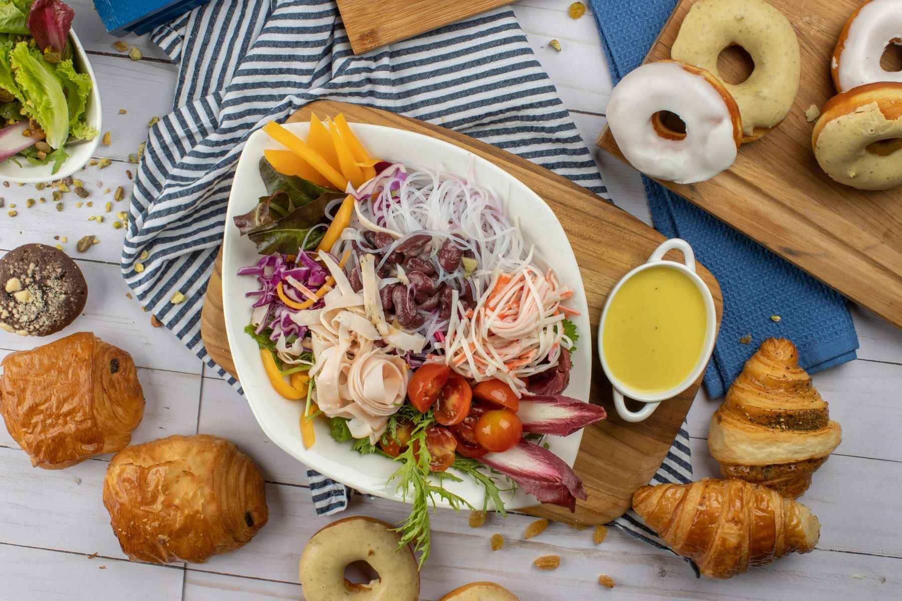 A colorful salad with various vegetables, cold cuts, and noodles is served in a white bowl with a small dish of yellow dressing. Surrounding the salad are croissants, pastries, donuts, and a muffin on a striped cloth.