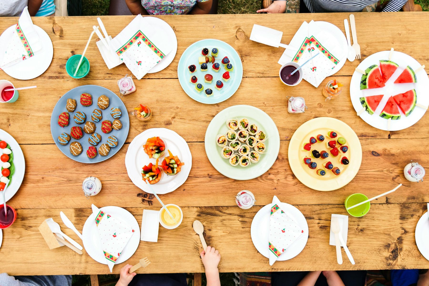 A wooden table set for a picnic with plates of colorful appetizers, watermelon slices, drinks, napkins, and cutlery. Several people sit around the table, visible only by their hands and arms.