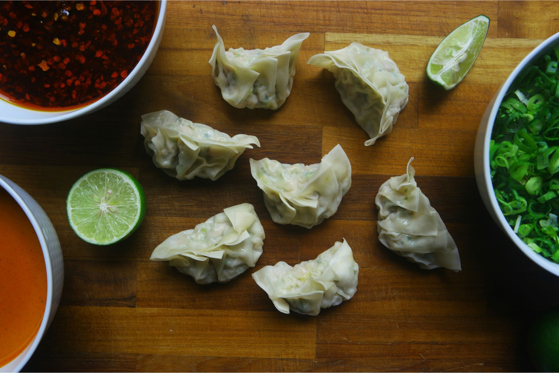 Seven dumplings are arranged on a wooden surface, surrounded by bowls of red and orange sauces, a bowl of chopped green onions, and two halved limes.
