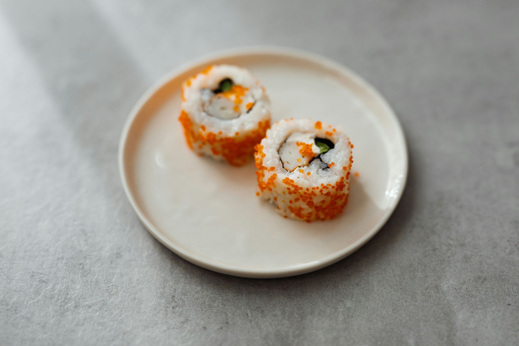 Two sushi rolls topped with orange fish roe are served on a small white plate, placed on a light gray surface.