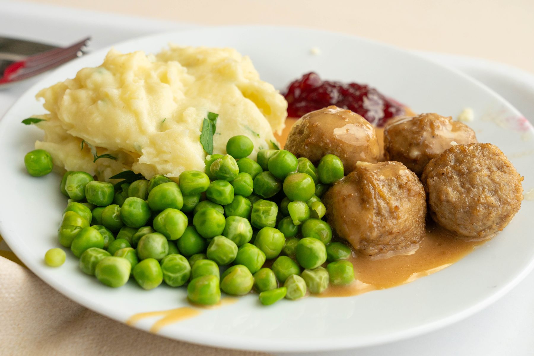 A plate with mashed potatoes, green peas, three meatballs in brown gravy, and a serving of red lingonberry sauce.