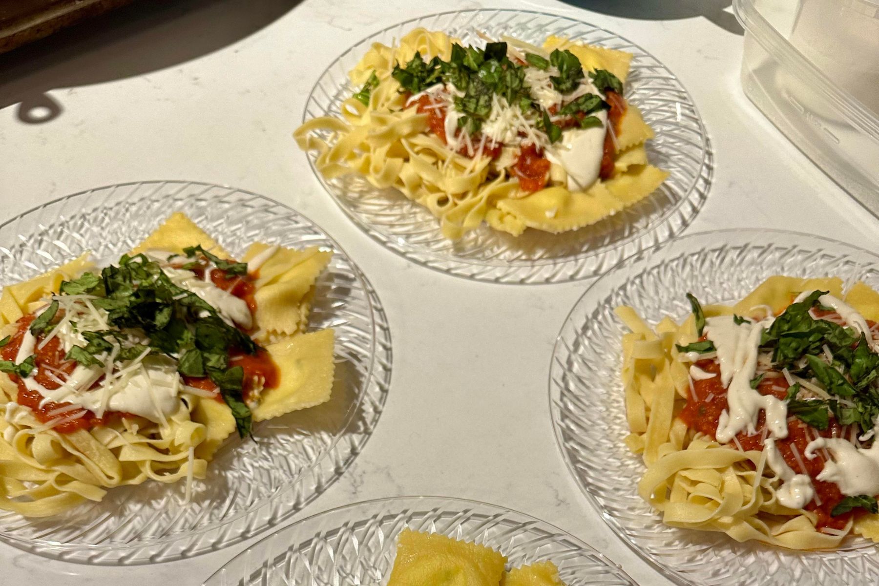 Three glass plates filled with pasta topped with tomato sauce, shredded cheese, chopped fresh basil, and a drizzle of white sauce, set on a light-colored tabletop.