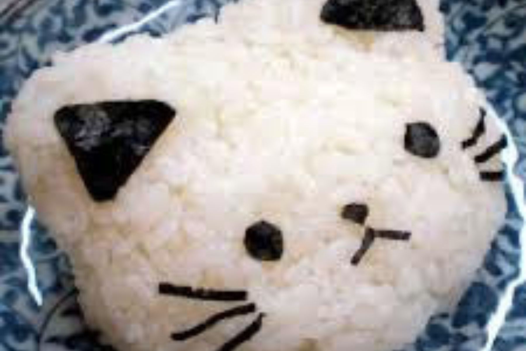 A rice ball shaped like a cat’s face, with seaweed pieces forming the ears, eyes, nose, and whiskers, placed on a blue patterned plate.