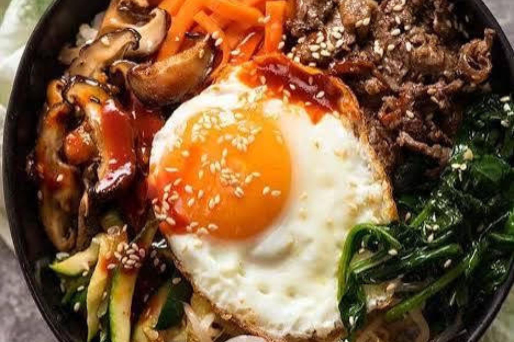 A bowl of bibimbap topped with a sunny-side-up egg, sautéed vegetables, beef, mushrooms, carrots, spinach, and sesame seeds, with rice underneath and a drizzle of sauce.