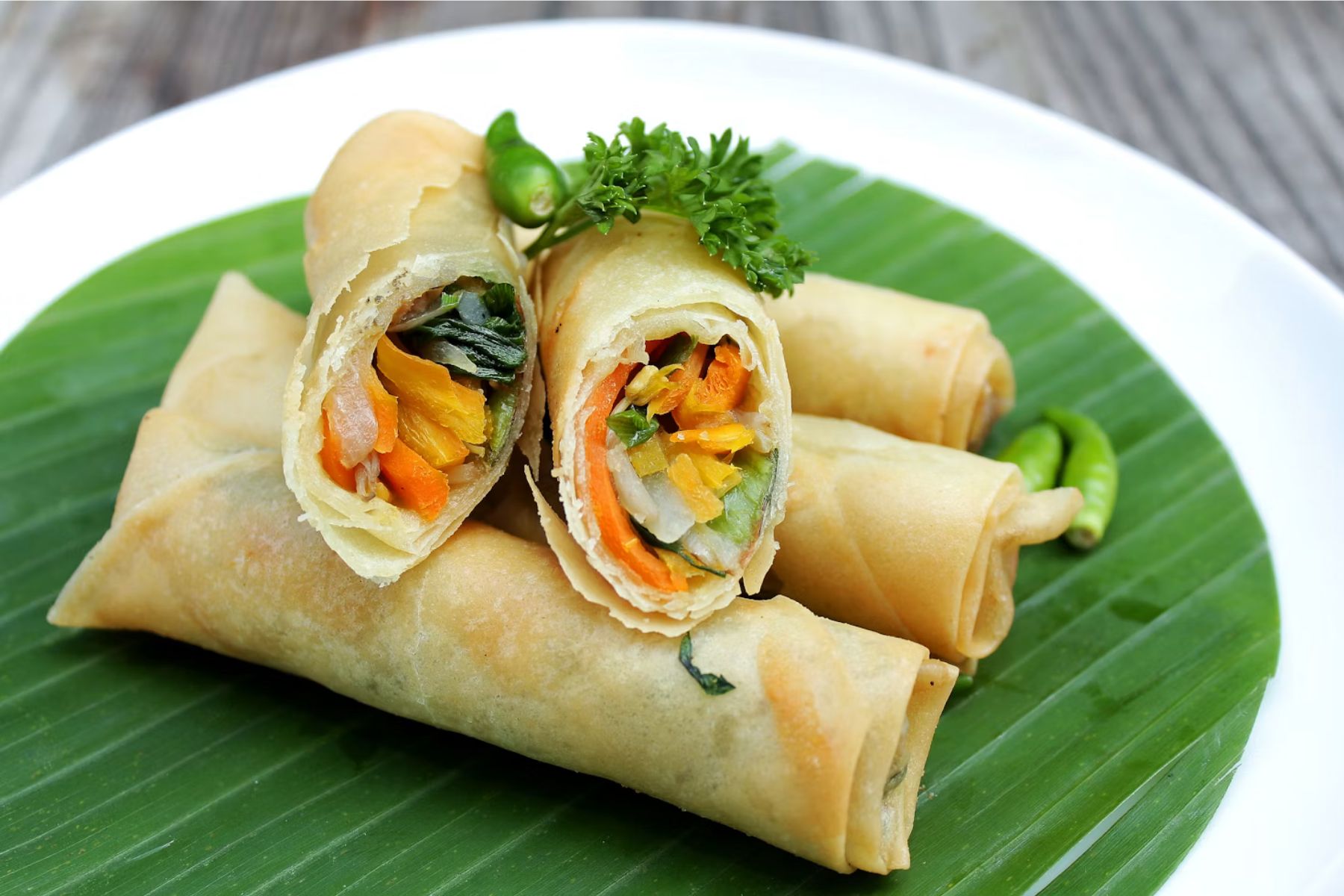 Three vegetable spring rolls are arranged on a banana leaf, with one cut open to reveal colorful vegetable filling. A sprig of parsley and green chili are used as garnish. The plate rests on a wooden surface.