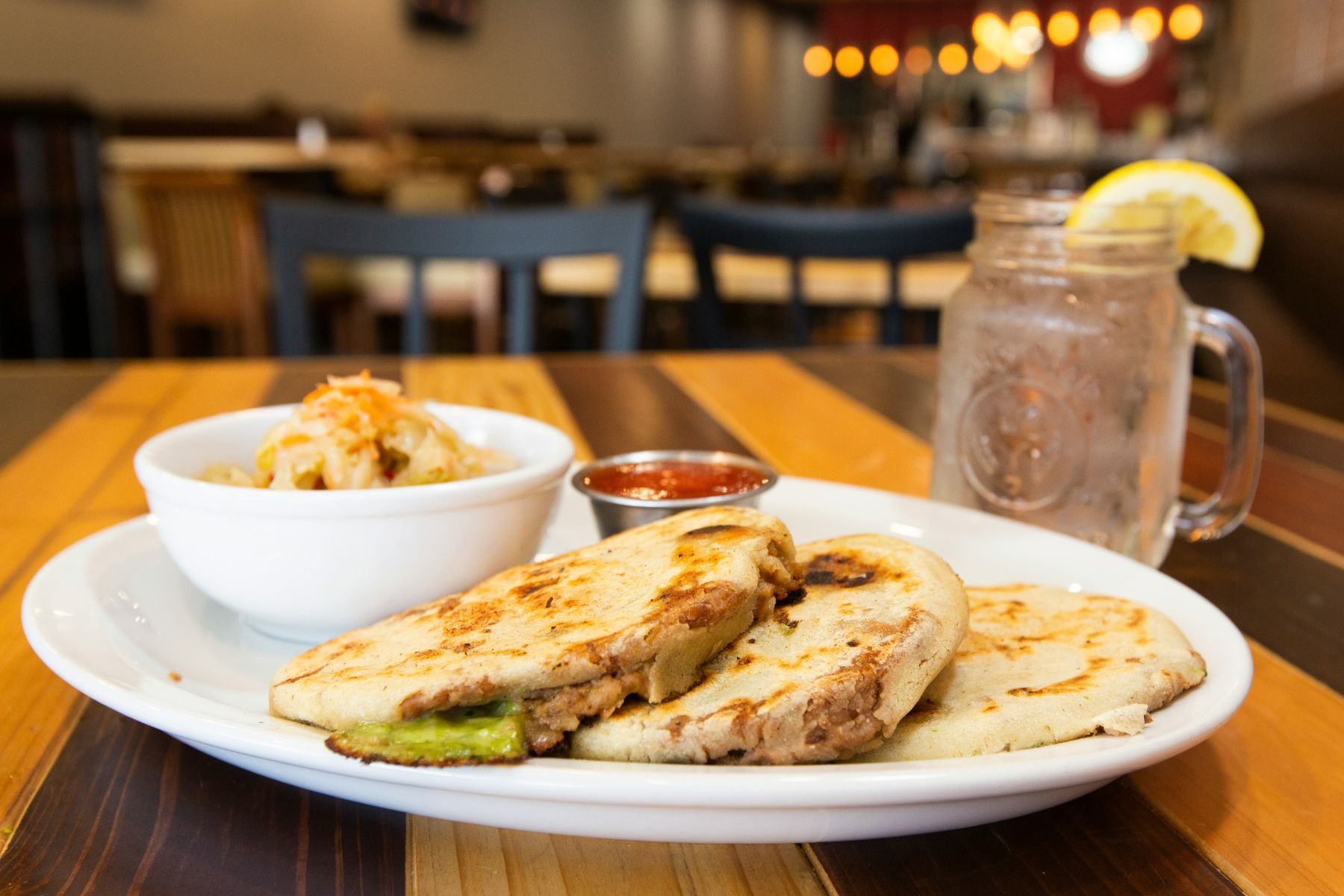 Two stuffed pupusas on a white plate with salsa, a bowl of pickled cabbage, and a mason jar of water with lemon on a wooden table in a restaurant setting.