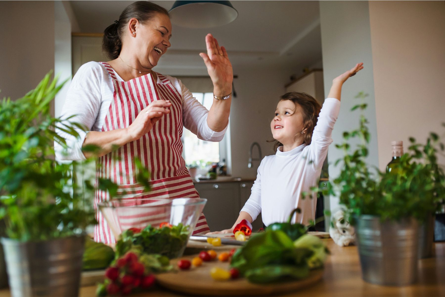 A woman and a young girl, surrounded by fresh vegetables in a kitchen, share a joyful high-five while preparing food together.