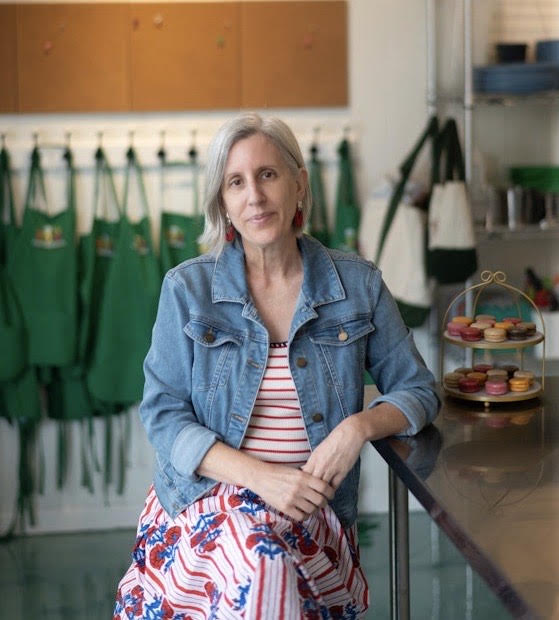 Meet the Chef - Charlene Kirk A woman with gray hair, wearing a denim jacket and a striped floral dress, sits at a kitchen counter with a tiered tray of macarons. Green aprons hang on the wall behind her.