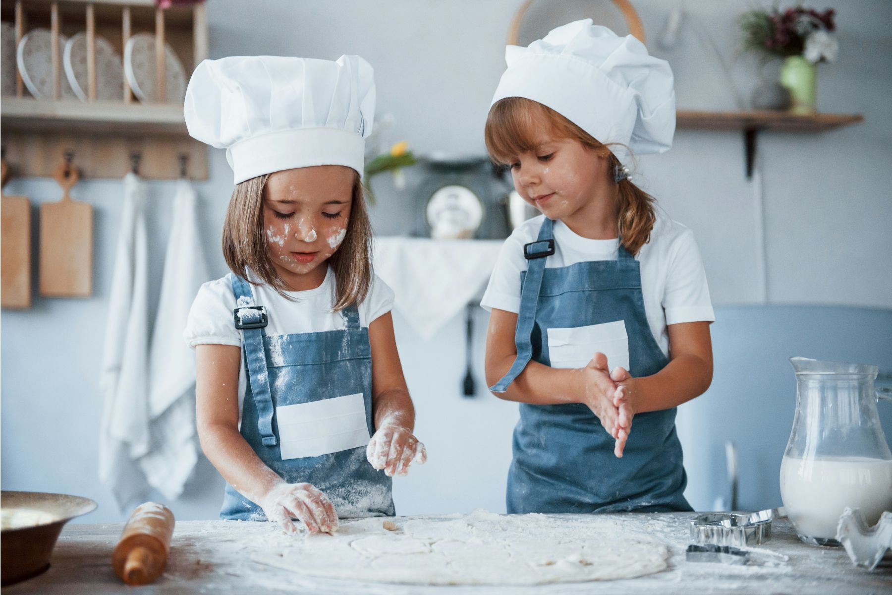 Two young girls wearing chef hats and aprons make dough together in a kitchen. Their faces and clothes have flour on them. A jug of milk, rolling pin, and cookie cutters are on the table.