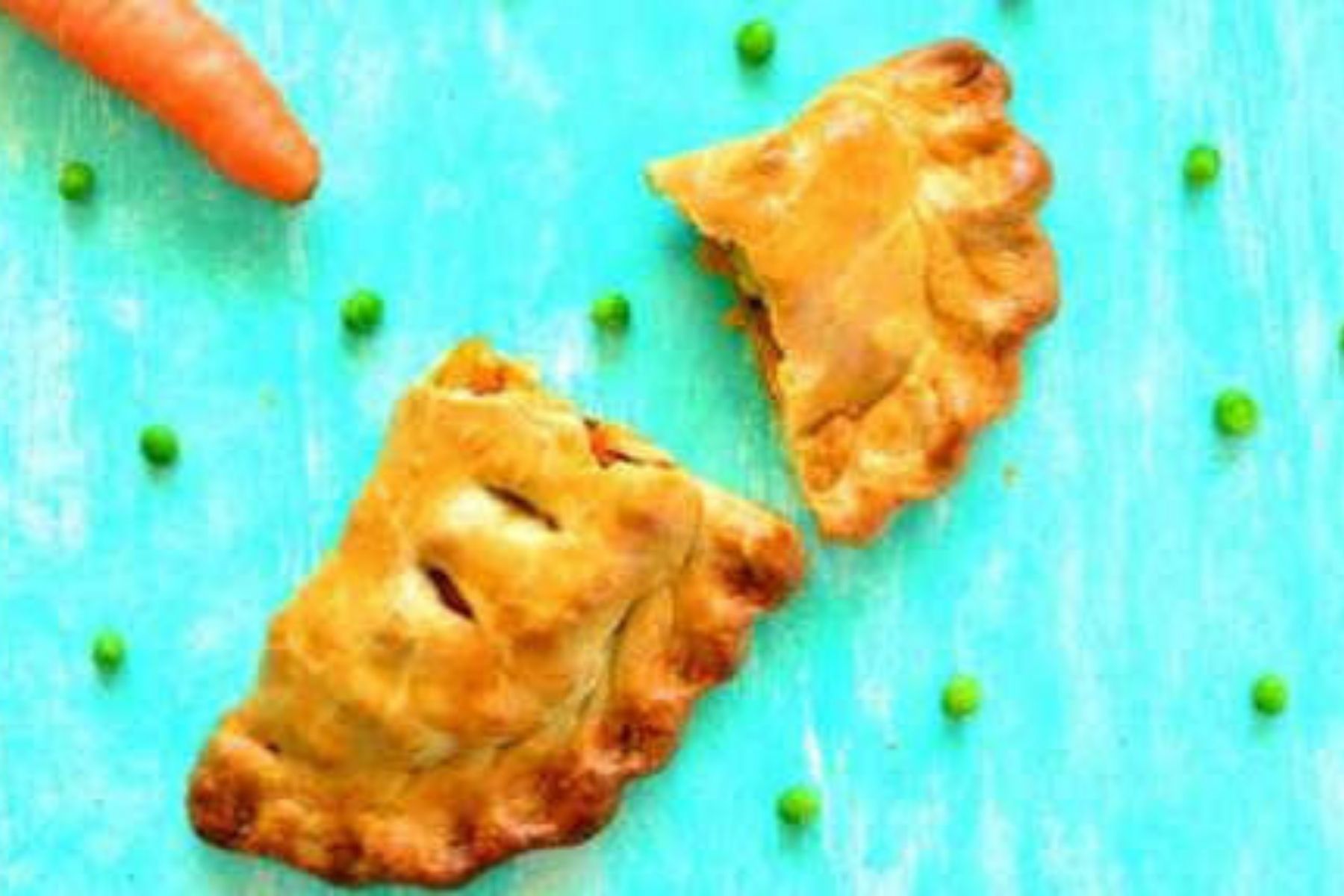 Two golden-brown baked pastries, one broken open, sit on a turquoise surface with scattered green peas and part of a carrot visible in the corner.