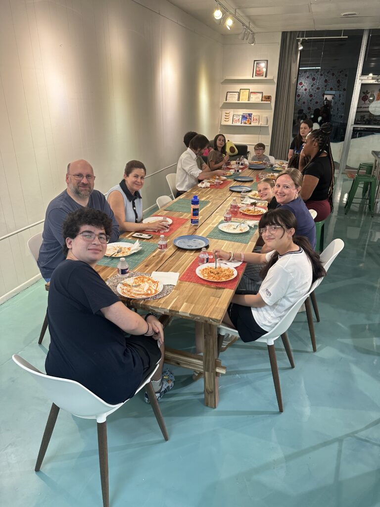 A large group of people sit around a long wooden table in a restaurant, eating pasta.