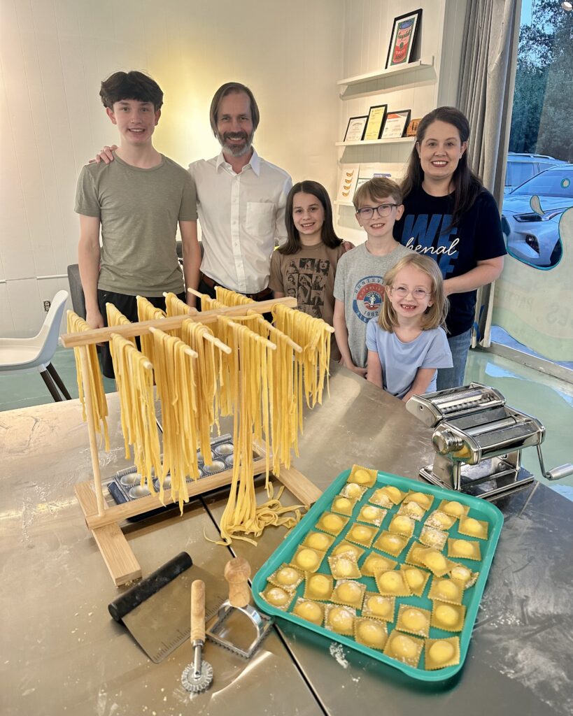 A family standing behind a stainless steel table with a wooden pasta drying rack filled with fresh noodles and a tray of prepared ravioli.