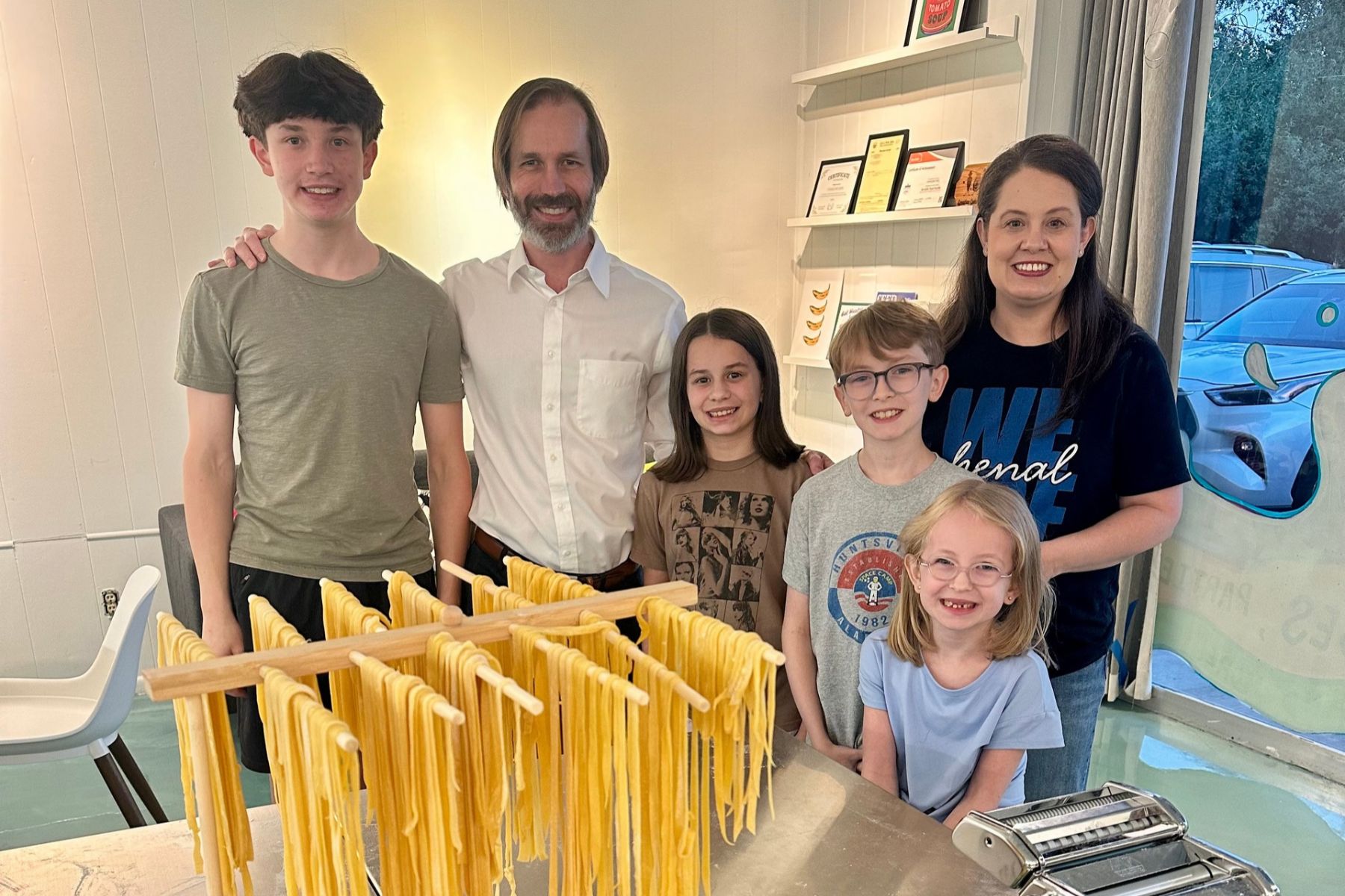 A family of six stands behind a wooden drying rack filled with fresh, hanging pasta noodles.