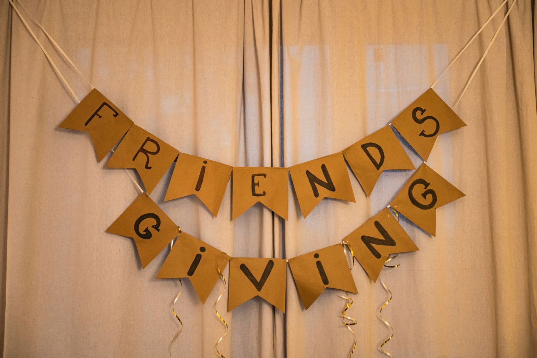 A two-tier brown paper banner spelling Friendsgiving hanging against a curtain.