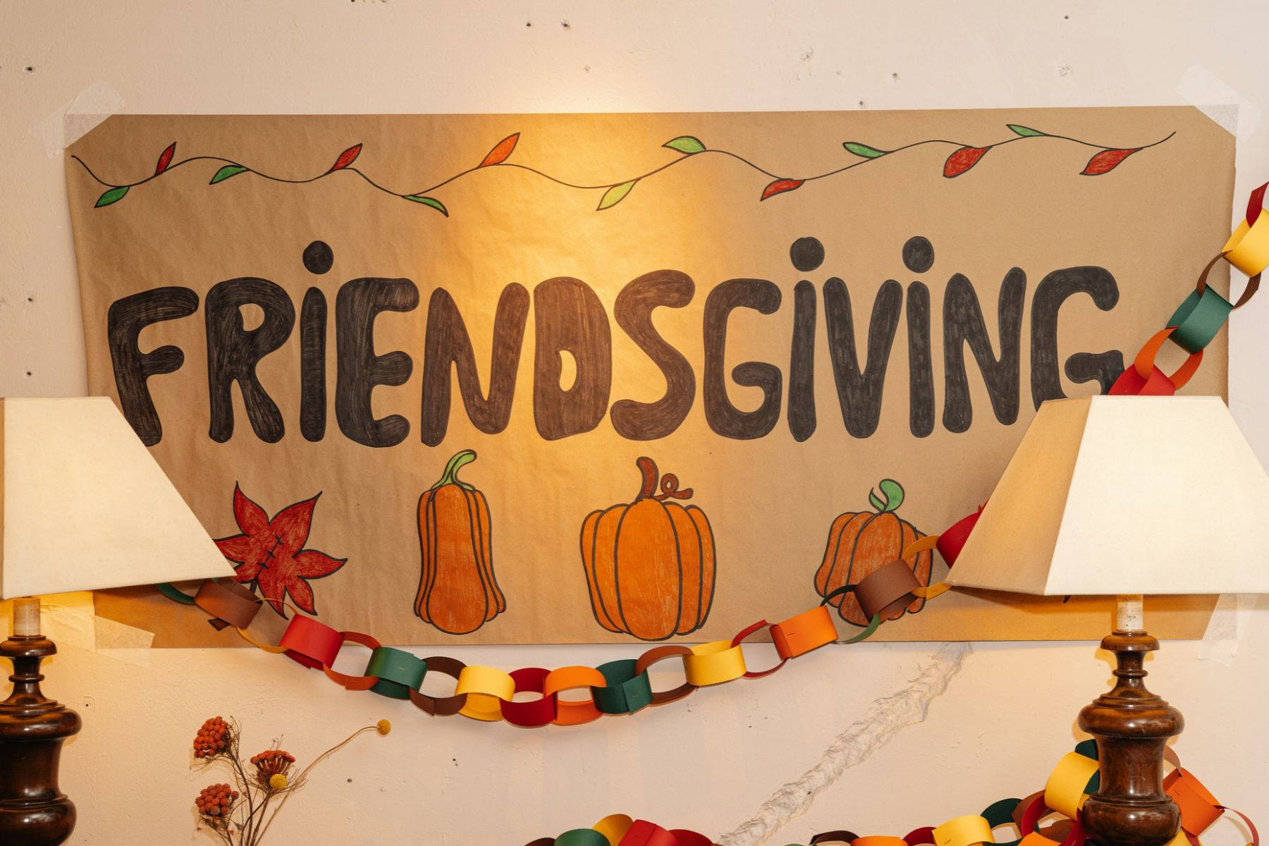 Hand-drawn FRIENDSGIVING sign on brown paper, decorated with illustrations of pumpkins and autumn leaves, draped with a paper chain garland.
