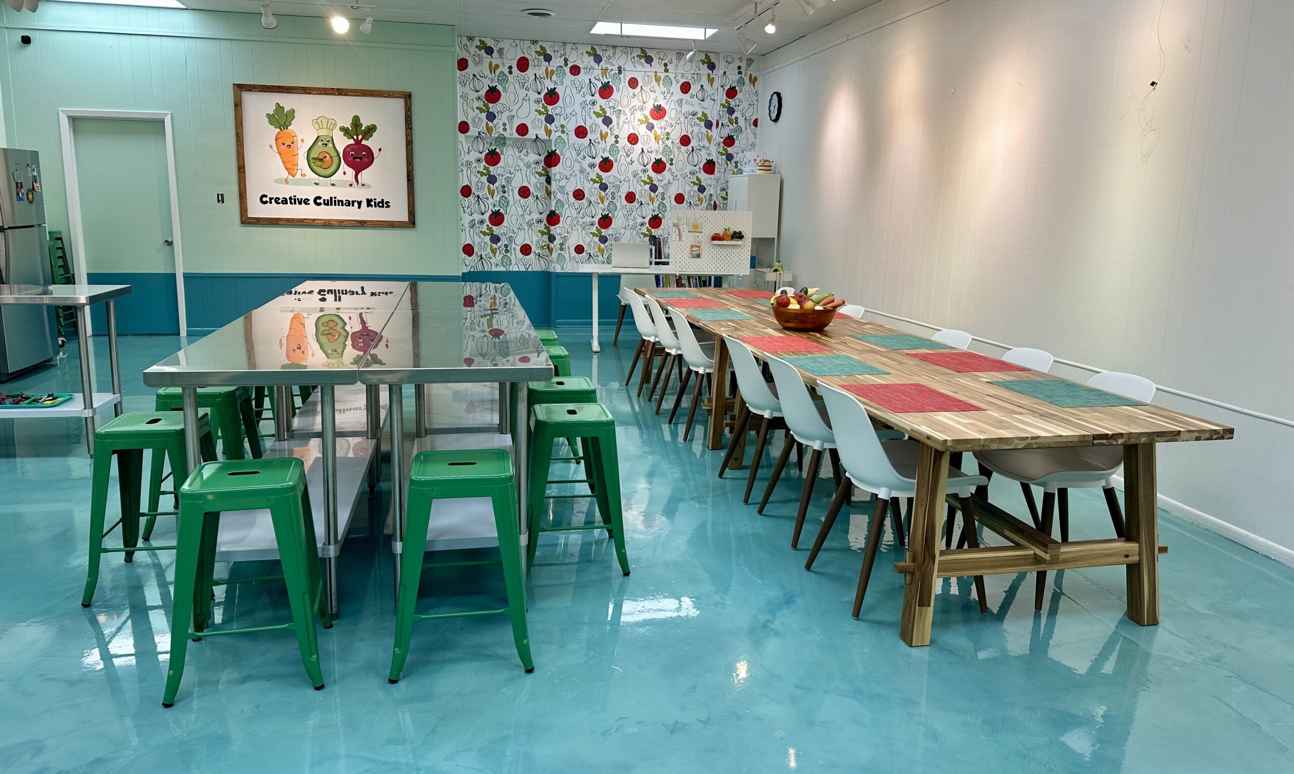 IMG_2851 A brightly lit culinary classroom for kids cooking classes, with a long stainless steel table and green stools on the left, a wooden dining table with white chairs on the right, and a Creative Culinary Kids sign on the wall.