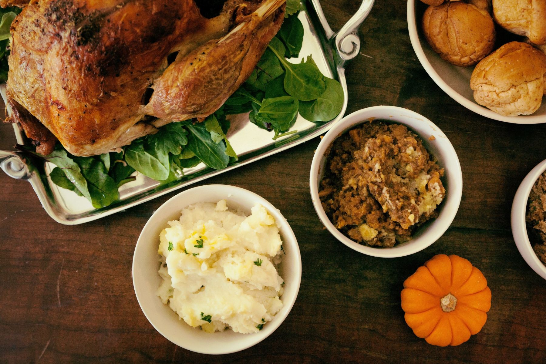 A roasted turkey on a bed of greens, served with bowls of mashed potatoes, stuffing, and dinner rolls on a wooden table with a small pumpkin.